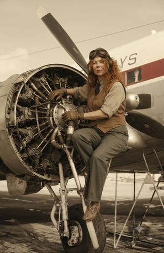 A vintage portrait of a red-haired aircraft mechanic seated beside an exposed radial engine and propeller.
